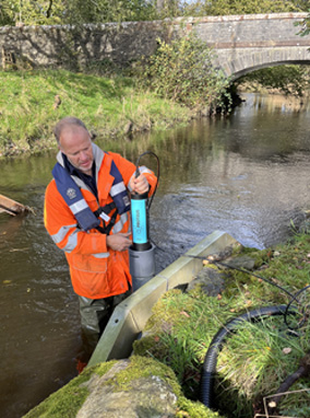 An engineer wearing a hi vis orange jacket stood in a river installing a Proteus in a stilling tube